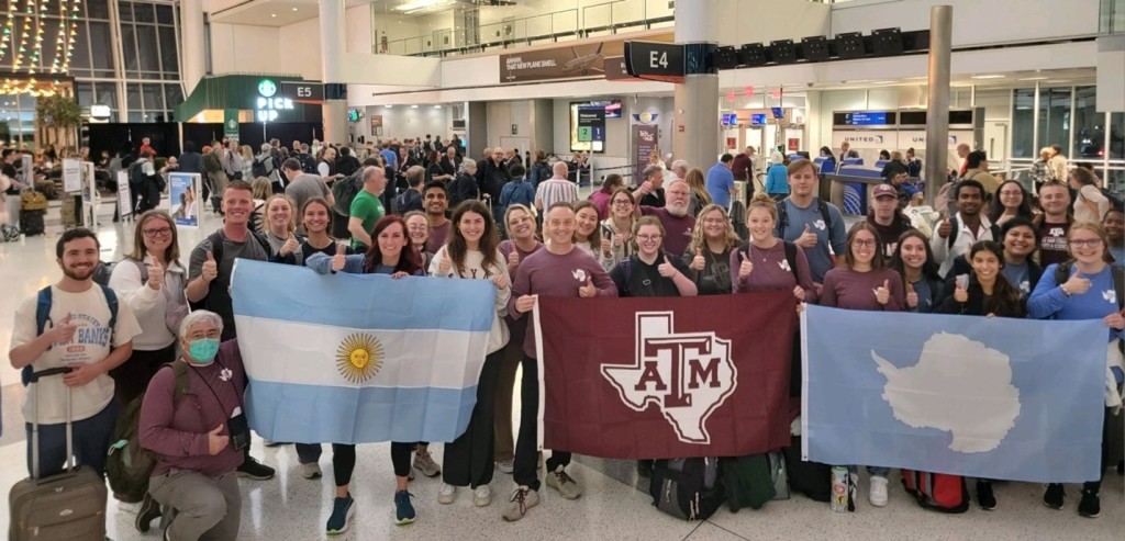 A group of people make thumbs-up gestures in an airport holding an Argentinian flag, a Texas A&M flag, and an Antartica flag.