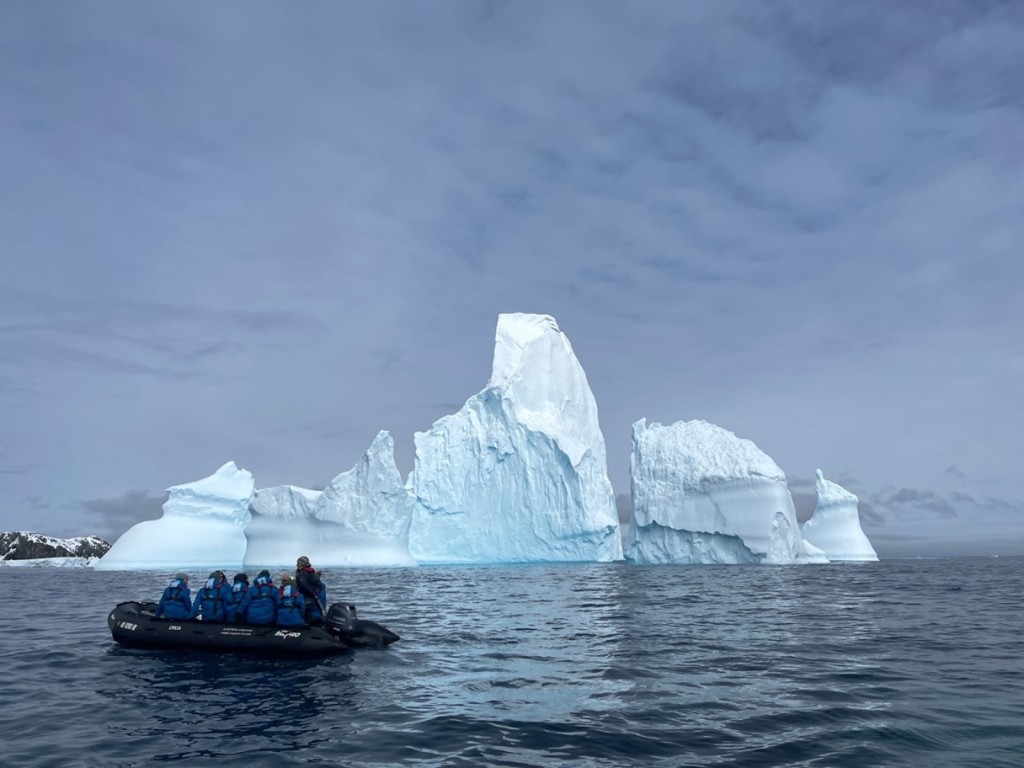 An inflatable boat filled with people in blue coats looking at an iceberg in the water